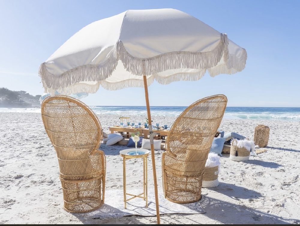 Fringed umbrella and peacock chairs set on white sand beside a styled beach table